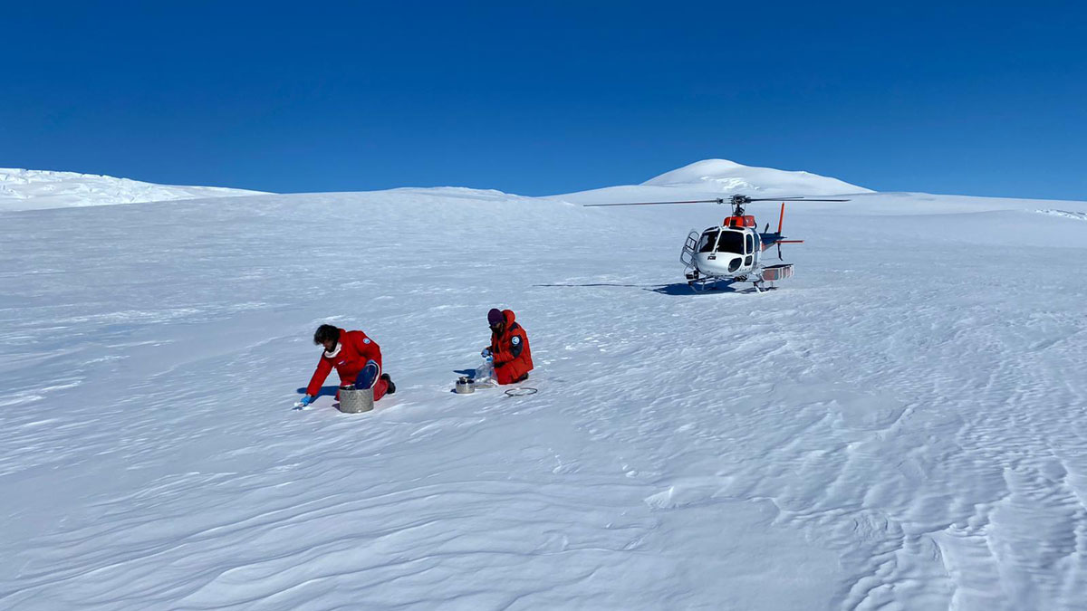 Two people in orange snowsuits kneel down in a snowy landscape in front of a stationary helicopter.