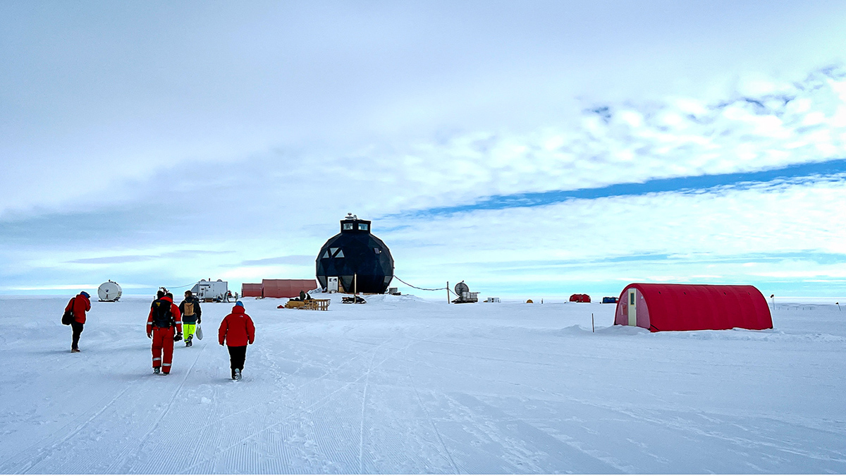 Researchers walk toward a large geodesic dome and a small red Quonset hut on the Greenland Ice Sheet.