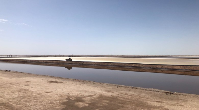 A long brine evaporation pond sits amid a bare, arid-looking landscape under a clear blue sky.