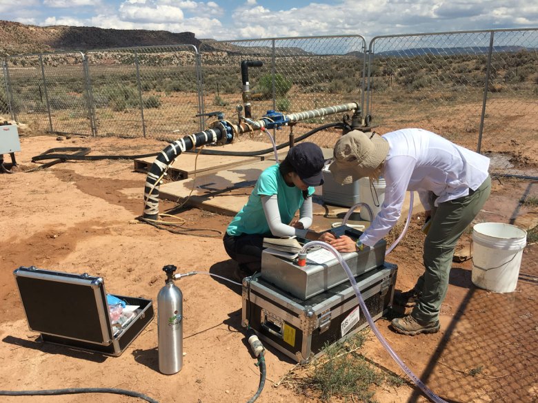 Two researchers huddle over groundwater sampling instrumentation in a fenced-in area amid red dirt and low scrubby vegetation.