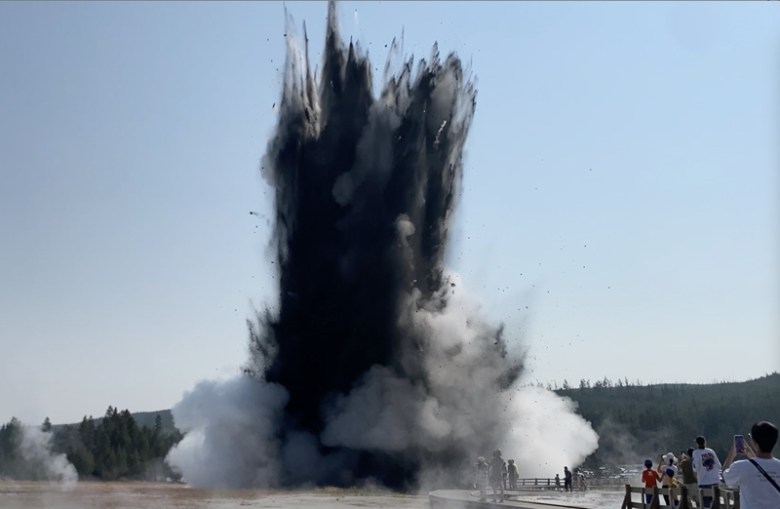 A tall plume of dark debris and white steam erupts from the ground as people in the foreground look on.