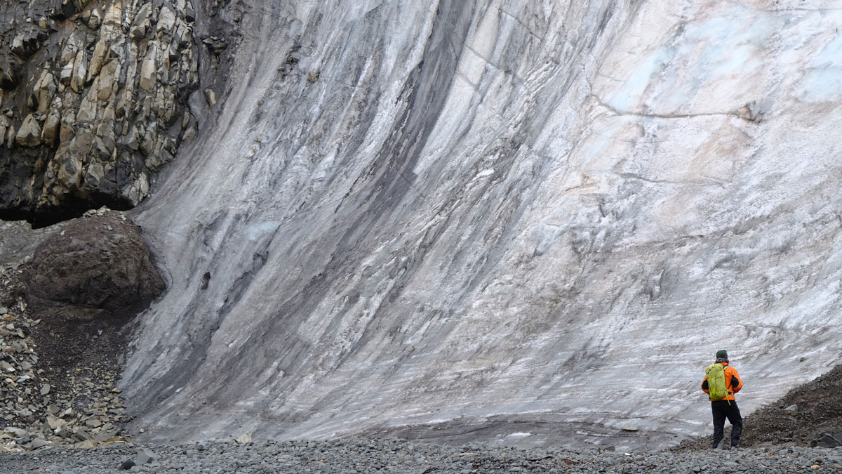 Tall glacier face with layered ice wall meeting rocky terrain, with a person in bright-colored clothing at the base.