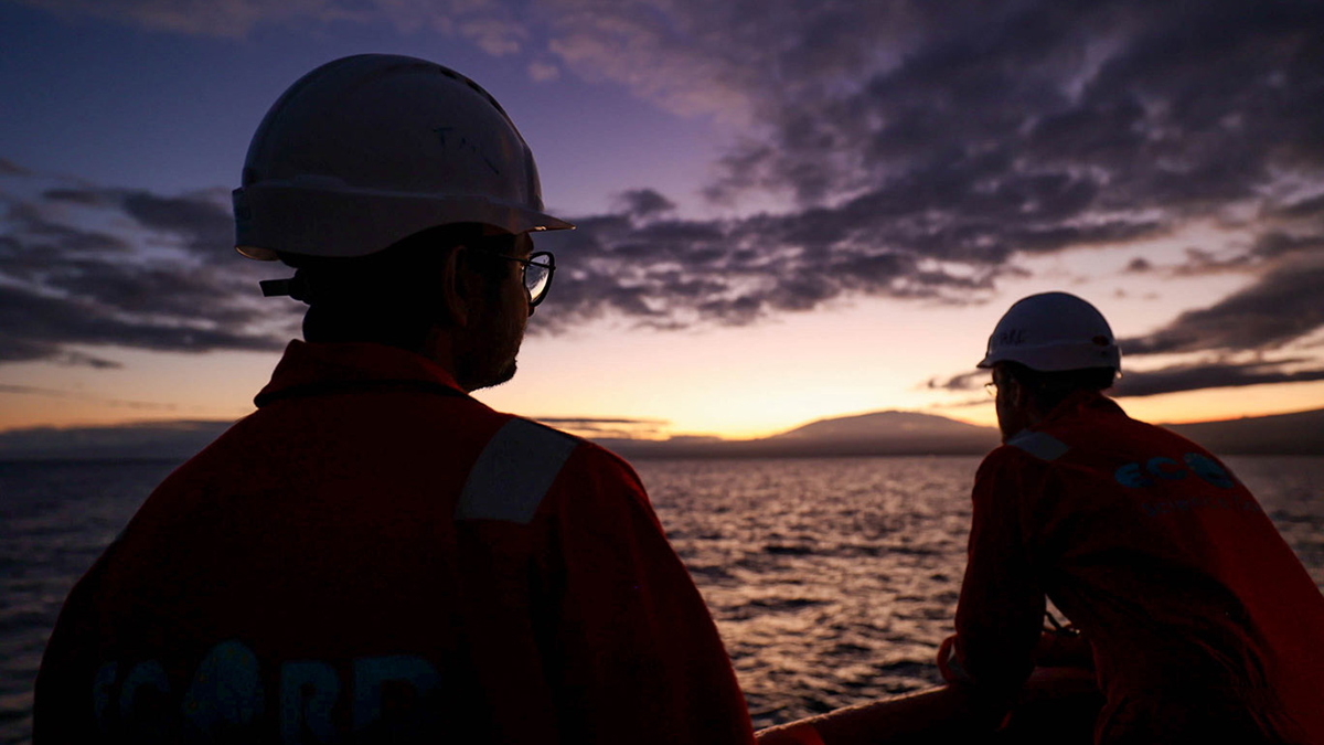 Two people wearing hard hats are silhouetted by the Sun rise in the distance as they look out over the ocean from a research vessel.