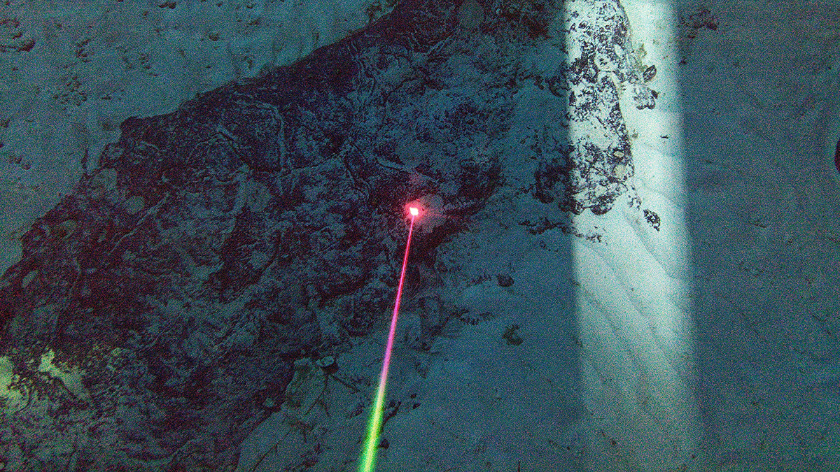 A narrow laser beam illuminates a point on a rocky seafloor outcrop surrounded by sand.