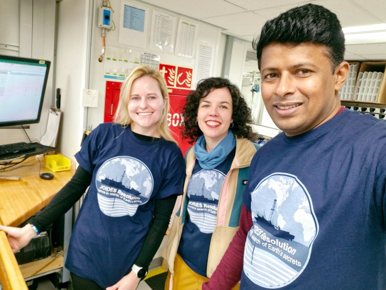 Three scientists garbed in JOIDES Resolution T-shirts smile at the camera.