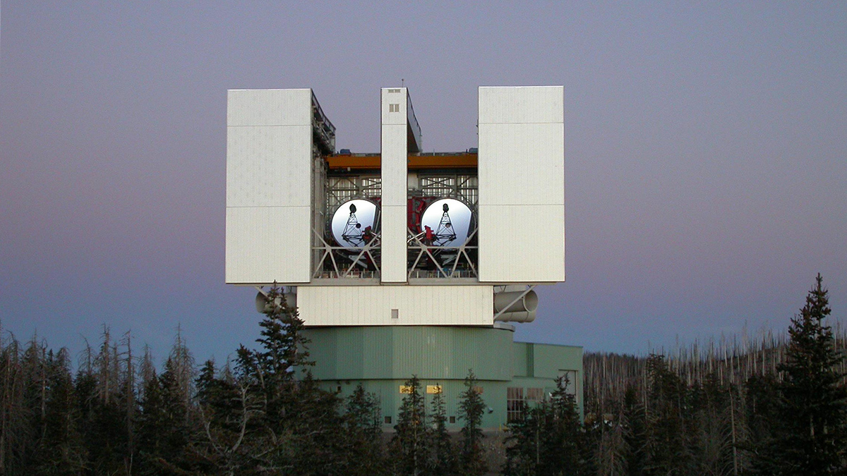 A mostly rectangular, man-made structure several stories tall sits among trees along a mountain ridge, against a dim sky. The structure is mostly white colored and has two large, round telescope dishes nested in between taller sections of the building and angled skyward.