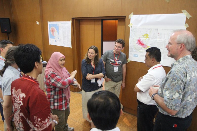A small group of people standing in a circle in a room with volcano hazard maps on the wall discuss the uses of such maps.
