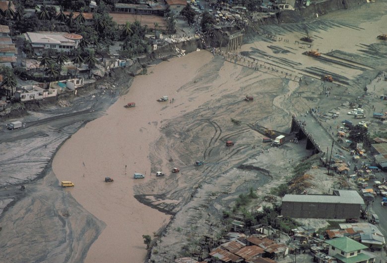 Aerial view of vehicles and people crossing a muddy river channel near where a bridge was washed out.