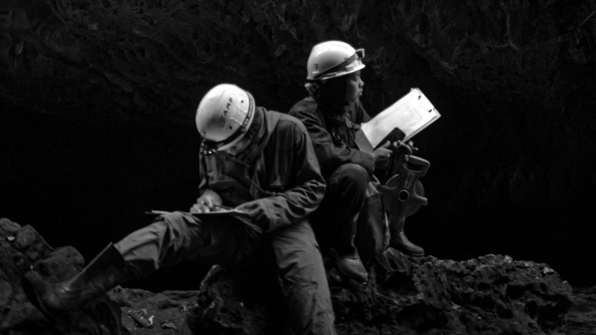 Black and white photo of people sitting down wearing helmets in a cave