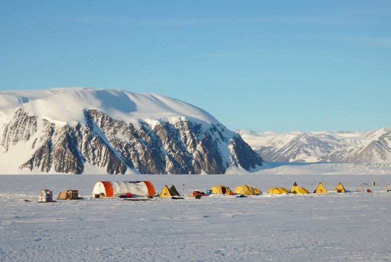 A line of mostly yellow tents at an Antarctic research site sits atop a flat, snow-covered expanse in front of imposing rocky bluffs.