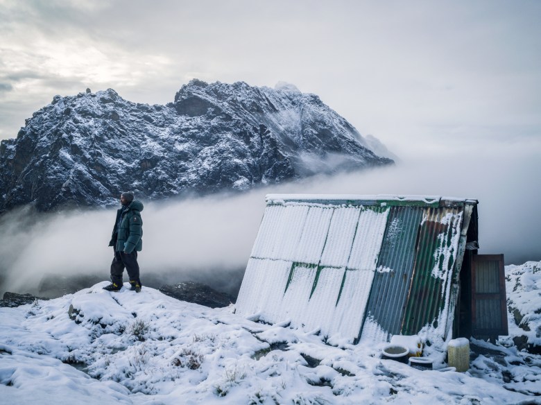 A man stands on a snowy mountain next to a boxy structure. He looks off into the distance at a peak.