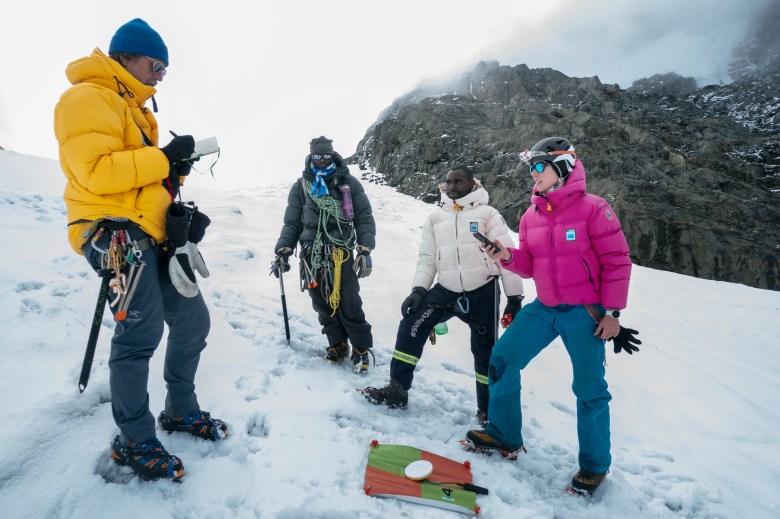 Four people stand on a snowy mountain around a green and orange square of fabric. They are wearing climbing gear and warm jackets.