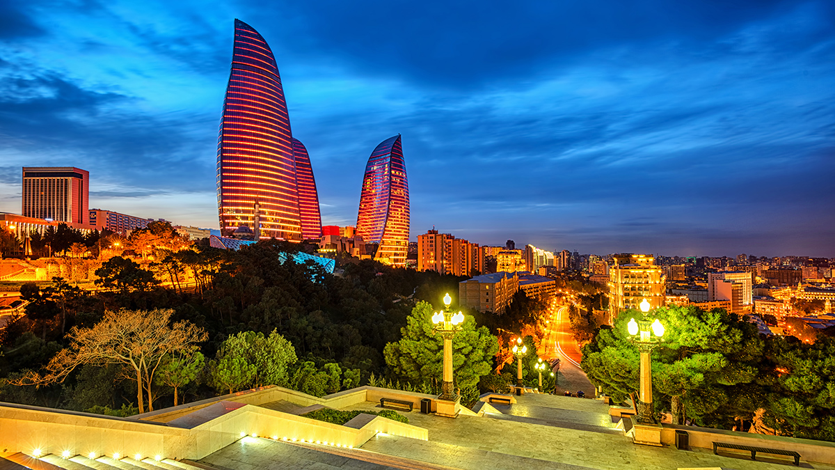 The Flame Towers, three skycrapers shaped like flames to symbolize fire, rise above other buildings against a deep blue evening sky. Bright lights shine from the buildings’ windows. In the foreground are treetops and street lights, and streaks of light from the headlights of moving traffic can be seen along a road below.