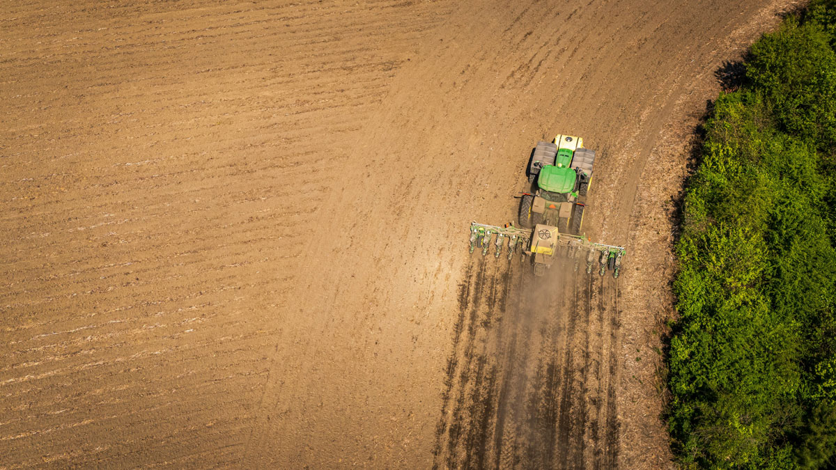 A tractor pulling a plow over a dirt field and generating dust is seen from above.