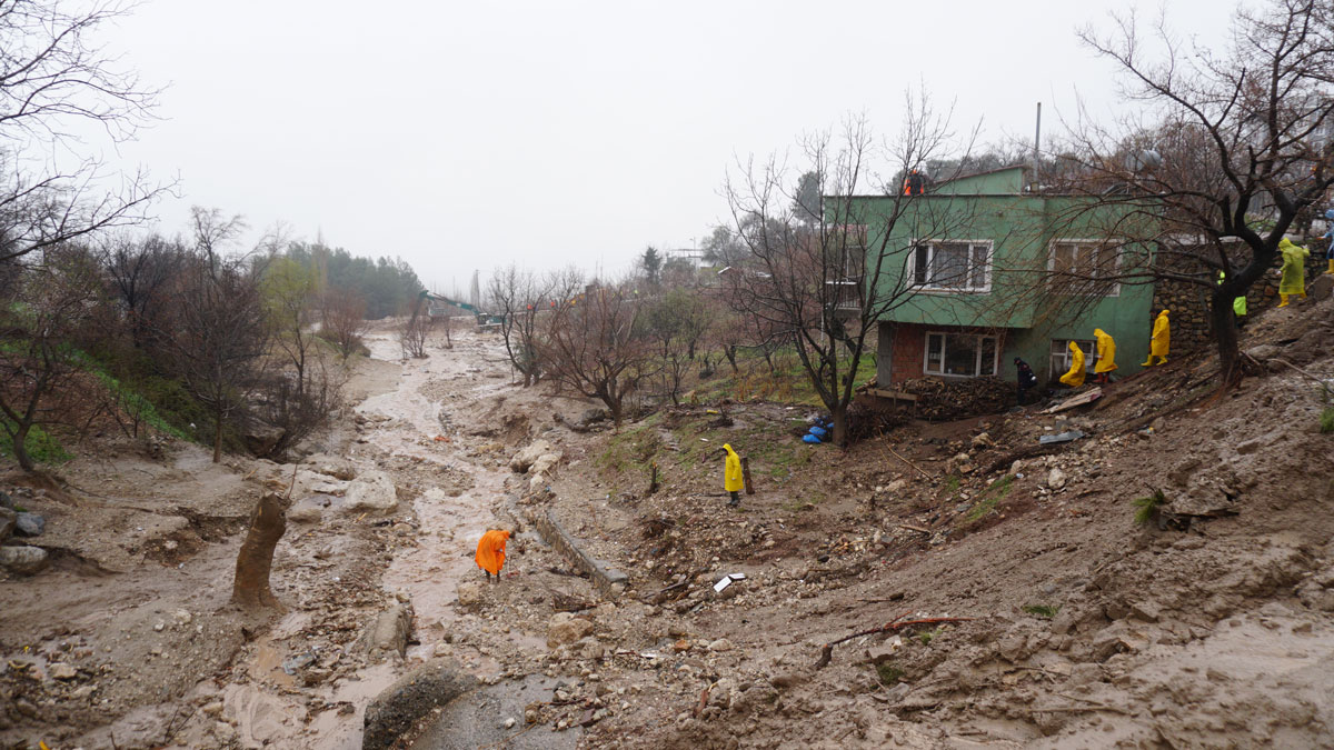 Muddy floodwaters flow through a ravine. People in bright orange and yellow raincoats assess damage near a green house on an eroded hillside.