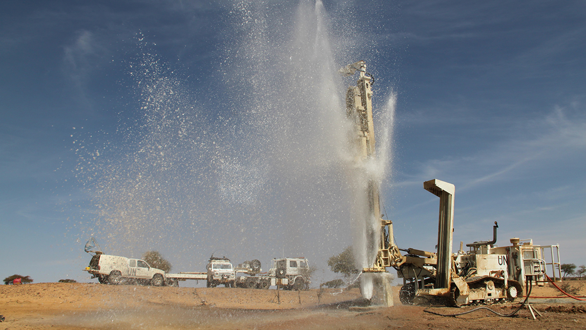 A plume water spouts high into the air from a well drilled in arid-looking ground beside water trucks and drilling equipment emblazoned with “UN.”