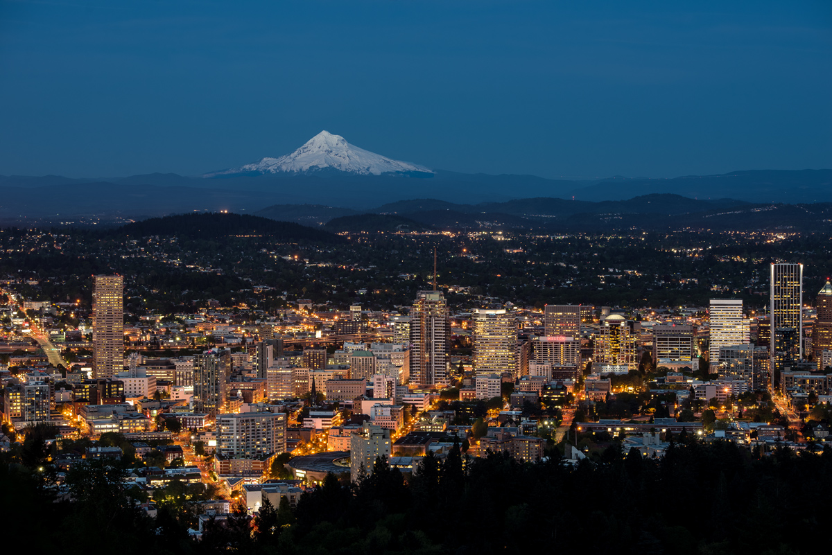 Snow-covered Mount Hood rises in the distance beyond the nighttime skyline of Portland, Ore.